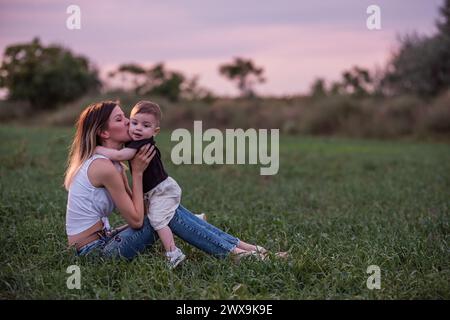 Smiling woman lifts, kiss little boy high in grassy field during serene sunset. Concept of pure joy, bonding, family connection. Mother and son walks Stock Photo