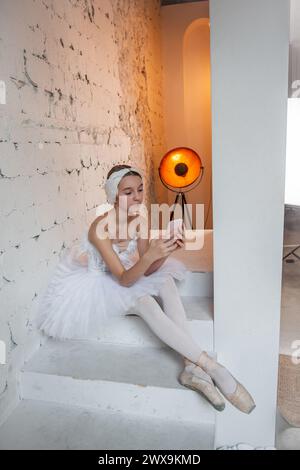Young ballerina in white tutu, with feather tiara, sits on the steps in loft interior, holds smartphone in hands, surfs the Internet. Teenager social Stock Photo