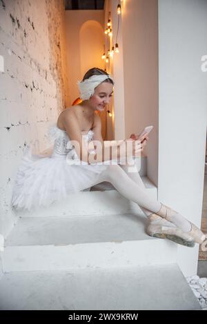 Young ballerina in white tutu, with feather tiara, sits on the steps in loft interior, holds smartphone in hands, surfs the Internet. Teenager social Stock Photo
