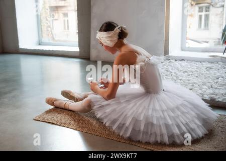 Young ballerina in white tutu, with feather tiara, sits on the floor in loft interior, holds smartphone in hands, surfing the Internet. Teenage social Stock Photo