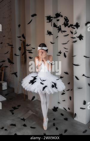 Dramatic young ballerina in white tutu, tiara stands on pointe, overwhelmed by falling black feathers, depicting scene of graceful chaos, delicate dis Stock Photo