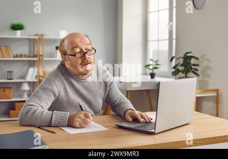 Senior man in eyeglasses using laptop and making notes on paper Stock Photo