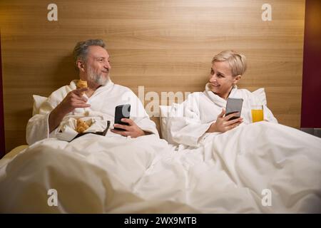 Smiling adult couple using smartphones and looking at each other on bed Stock Photo
