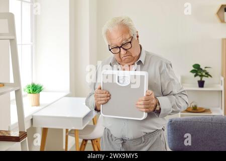 Upset retired grey-haired senior man holding weighing machine standing in the living room at home. Stock Photo