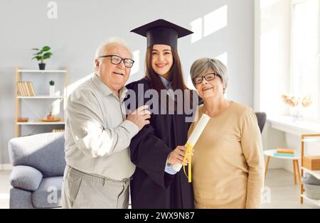 Young graduate girl holding diploma standing with senior old parents proud of her at home. Stock Photo