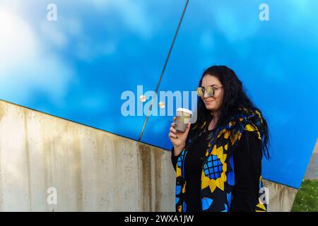 Beautiful young brunette woman with long curly hair, dressed in a black, blue and yellow waistcoat, drinks coffee from a paper cup on a background of Stock Photo