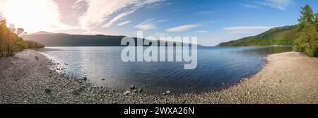Panoramic view across Loch Ness, Scotland, from a curving pebble shore, with rippled water, wooded hills and low sunlight beneath a blue sky. Stock Photo