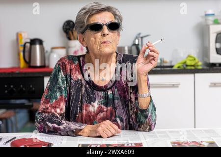 Portrait of old smoker sitting in her modern white kitchen with dark sunglasses. A woman from another time. Stock Photo