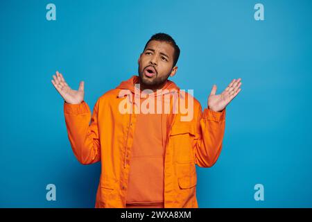 confused african american man in orange outfit putting hands to sideways on blue background Stock Photo