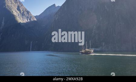 Boat sailing through fjord with steep rocky walls and waterfall in distance, Fiordland, New Zealand. Stock Photo