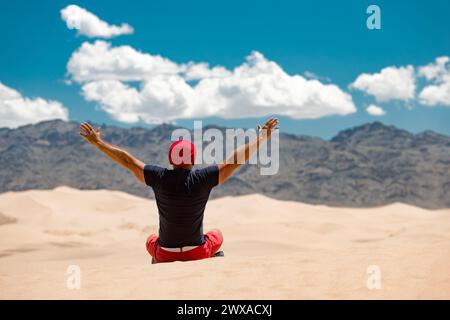 Happy tourist in red cap is sitting with open arms against sand dunes, mountains and blue sky. Travel in desert concept Stock Photo