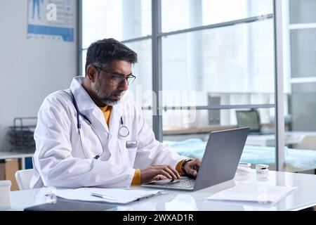 Portrait of senior Middle Eastern doctor using laptop while sitting at desk in medical clinic copy space Stock Photo