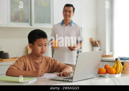 Vietnamese schoolboy watching educational video on laptop and doing homework Stock Photo