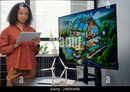 Screen with new architectural project and two models of windmill standing on desk against young female employee making presentation Stock Photo