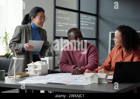 Three happy young intercultural architects discussing new project while one of them pointing at unrolled blueprint with sketch of house Stock Photo