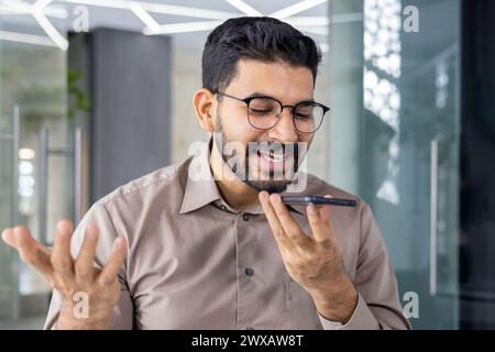 Smiling young adult male engages in a cheerful conversation using a hands-free mobile phone in a modern office setting. Stock Photo