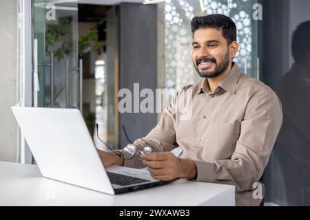 Happy young businessman working on laptop at desk in contemporary office setting, showing job satisfaction and professionalism. Stock Photo