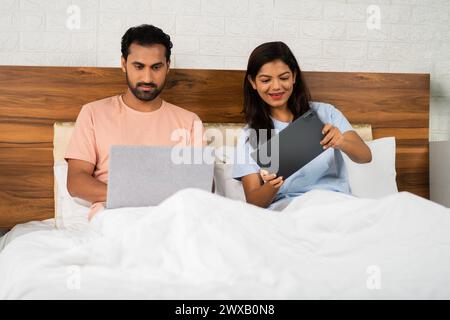 Husband working on laptop while playing game on tablet on bedroom while sleeping at night - concept of technology, digital gadgets and relationship Stock Photo