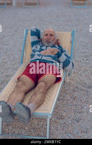 An old man is lying on a sun lounger on the beach. An elderly gray-haired tourist with a beard in red shorts is enjoying himself on the beach by the s Stock Photo