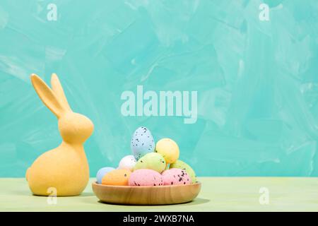 Easter greeting card. Yellow Easter bunny and pile of decorative colorful eggs in round wooden plate. Turquoise white background as space for text. Ho Stock Photo