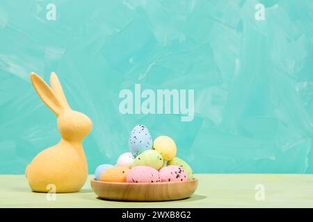 Easter greeting card. Yellow Easter bunny and pile of decorative colorful eggs in round wooden plate. Turquoise white background as space for text. Ho Stock Photo