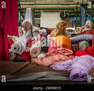 Muslim ladies wearing head scarfs buying cloth and material from rolls of material on an open air market stall Stock Photo