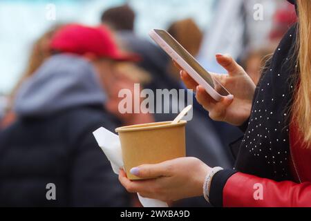 Womans hands holding bowl of food and mobile phone, midsection of womans body taking selfie of a meal at farmers street food market at Prague Naplavka Stock Photo