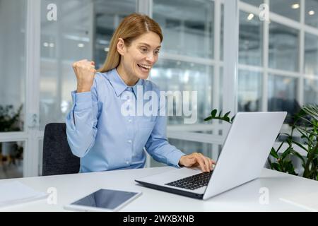 A joyful businesswoman is celebrating a successful moment while working on her laptop in a modern office setting. Stock Photo