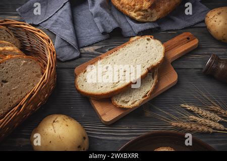 Freshly sliced rustic bread on a cutting board, with potatoes and wheat, on a dark wood surface Stock Photo