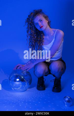 Woman with disco ball is striking pose in squatting position in blue neon colored lighting during celebration party in nightclub Stock Photo