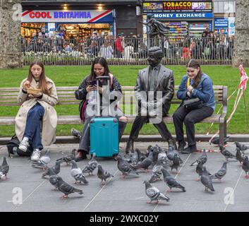 Women sitting on a bench next to a bronze statue of Mr Bean, in Leicester Square, London. Pigeons gather in front of them in search of food. Stock Photo
