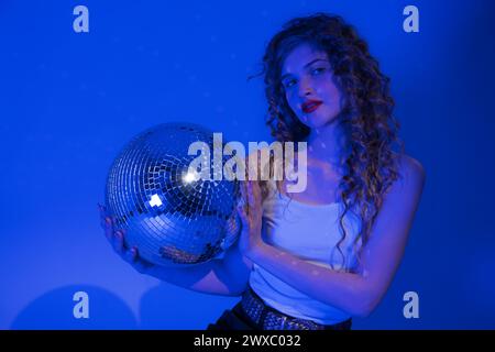Woman looks absolutely stunning as she poses with disco ball in nightclub. Blue neon lights add touch of sensuality to scene Stock Photo