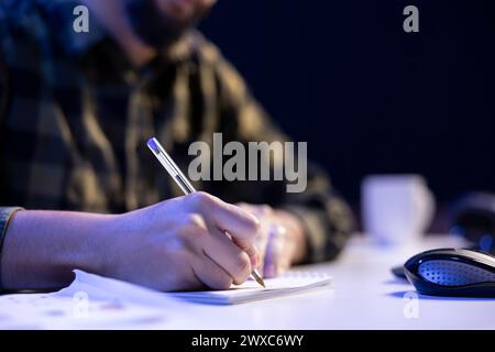 Person studying and researching information while writing on a notepad at a table. Closeup of male individual making notes with a pen on paper, organizing their schedule, and finishing tasks. Stock Photo