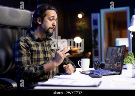 Male student having virtual conversation with classmates, engrossed in online meeting. Young man checking his notes and utilizing his laptop for effective communication through video conferencing. Stock Photo