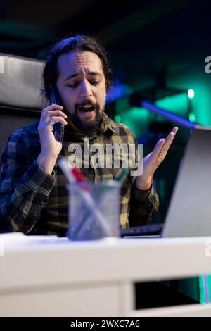 At desk, young man engages in an important conversation on his mobile device while checking his laptop. Dedicated male freelancer sitting at the table and speaking with clients on his smartphone. Stock Photo