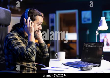 Male freelancer working at a desk focused on his laptop that is displaying code and data. Man wearing wireless headphones is checking software information and ensuring cyber security. Stock Photo