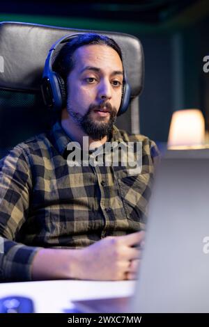 Male individual wearing wireless headphones and looking at his digital laptop. Sitting at a desk is young man utilizing his headset and portable computer for entertainment, watching a movie at home. Stock Photo