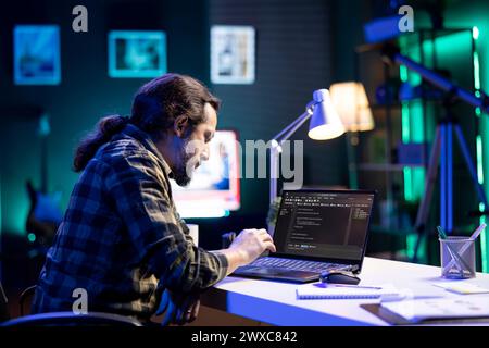 Dedicated male programmer uses his portable computer to manage databases and keep an eye on network security. Bearded man is coding on a digital laptop while seated at a desk. Stock Photo