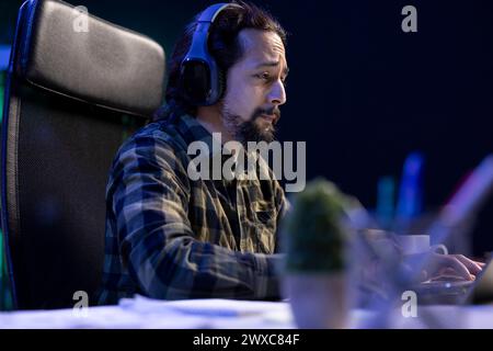 A focused man wearing a wireless headset attends a video call meeting with clients on his portable computer. A male with a sad expression works remotely, using a laptop and headphones. Stock Photo