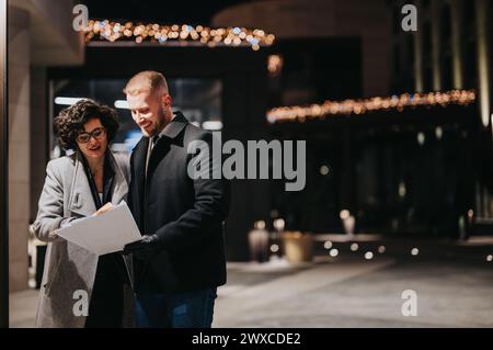 A business couple are smiling and engaging in a cheerful discussion over documents outside an illuminated building at night. Stock Photo