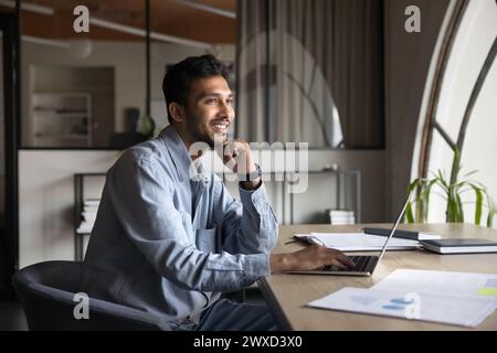 Happy dreamy young Indian professional man sitting at laptop Stock Photo