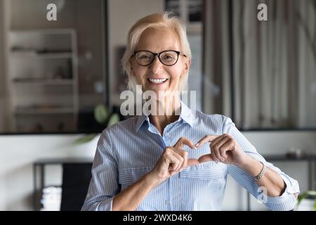 Cheerful senior blonde business lady making hand heart gesture Stock Photo