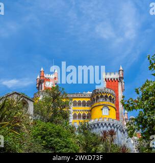 View from below of the Pena Palace in a colorful yellow and red style on fortified walls covered with greenery and palm trees under a sunny blue sky. Stock Photo