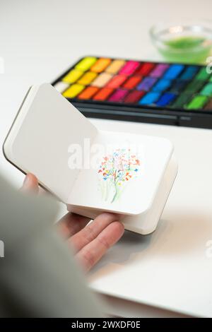 One hand shows a mini watercolor notebook with a miniature floral drawing with a colourful watercolour kit in the background Stock Photo