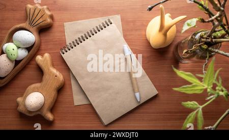 Blank craft paper notepad, pen among Easter decorations figured wooden plates in form bunny and carrot with eggs, twigs with spring leaves on wooden d Stock Photo