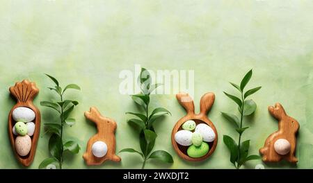 Row of wooden figured plates in form of bunnies and carrot with decorative Easter eggs and green ruskus twigs on textured green yellow background. Hol Stock Photo