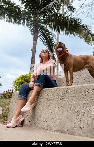 Middle-aged woman seated outdoors wearing red headphones and sunglasses, enjoying the sunny day with her pet. Stock Photo