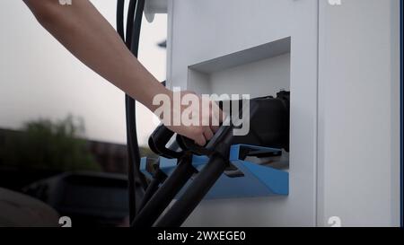 Hand holding EV charging plug in front of camera with blurred background of outdoor. Driver of electric car hold batteries charger plug. Sustainable e Stock Photo