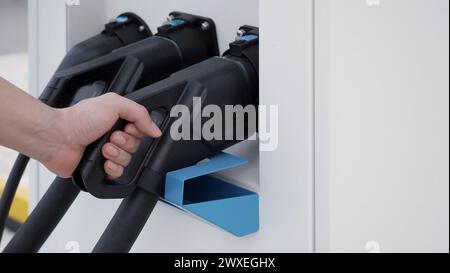 Hand holding EV charging plug in front of camera with blurred background of outdoor. Driver of electric car hold batteries charger plug. Sustainable e Stock Photo