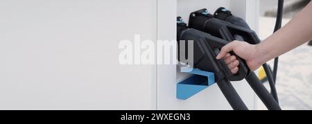 Hand holding EV charging plug in front of camera with blurred background of outdoor. Driver of electric car hold batteries charger plug. Sustainable e Stock Photo